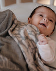Ein fröhliches Baby liegt zugedeckt mit einer personalisierten lilimoon Babydecke in hellgrau und cremeweiss mit Blütenmuster im Kinderbett.