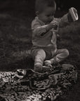 Ein kleiner Junge sitzt im Garten auf einer lilimoon Babydecke in schwarz und weiß mit Blütenmuster und spielt mit Holzspielzeug.