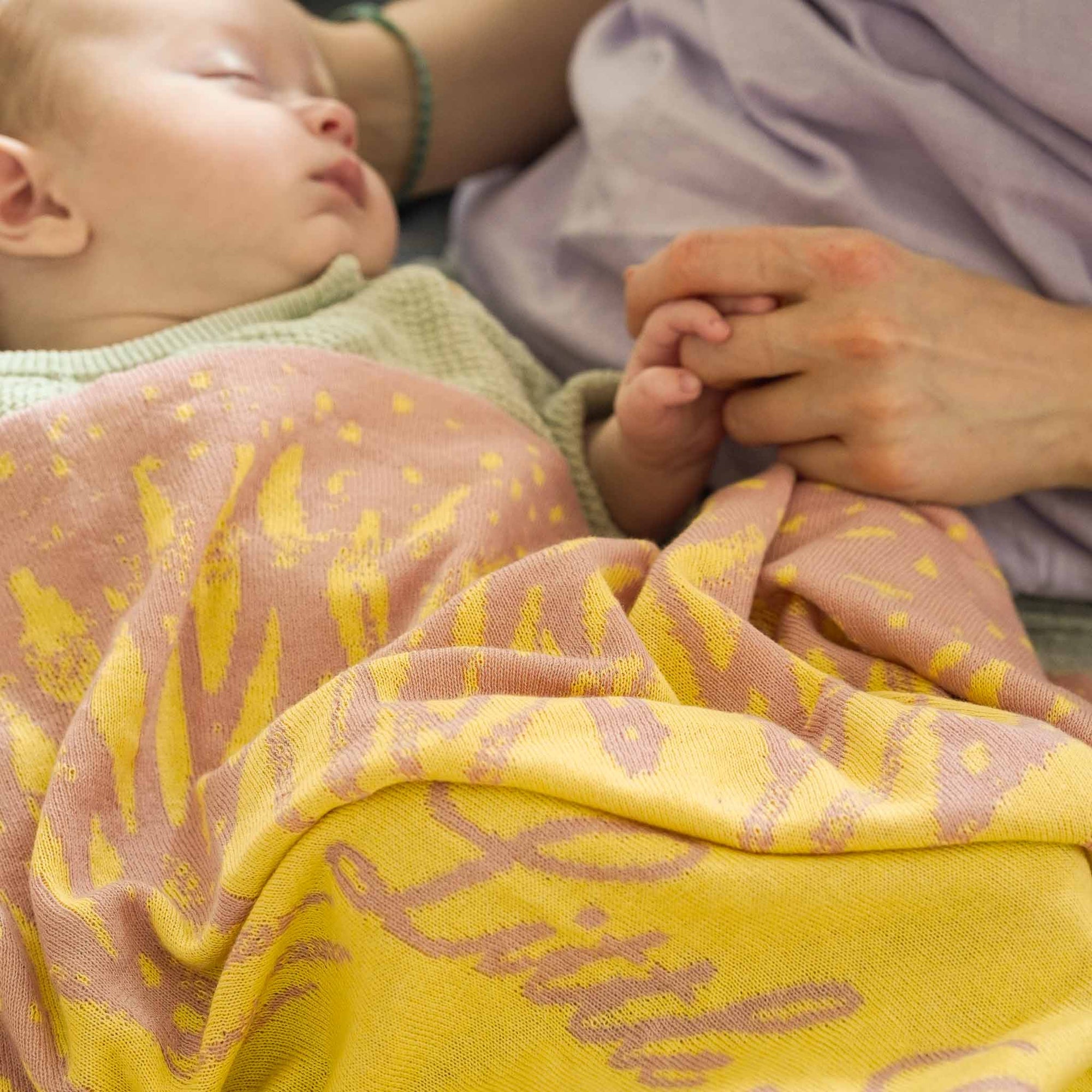 Baby schläft zugedeckt von einer sonnengelben Babydecke auf dem Schoß seiner Mama und sie hält seine Hand.