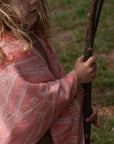 Kleinkind hat personalisierte lilimoon Babydecke gestrickt aus Baumwolle und Kaschmir in Korallenrosa und Cremeweiss wärmend um die Schultern gelegt und spielt mit einem Stock in der Natur.
