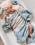 Ein neugeborenes Baby liegt kuschelig umhüllt von einer gestrickten lilimoon Babydecke in hellblau und cremeweiss mit Blütenmuster in einer Babywiege.