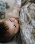 Ein Baby liegt zufrieden und zugedeckt mit einer kuscheligen lilimoon Babydecke in hellgrau und cremeweiss mit Blütenmuster in seinem Babybett.