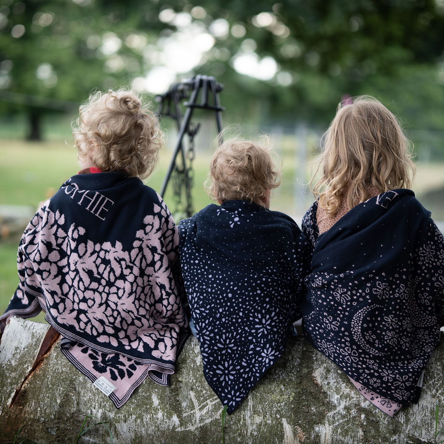 Drei Kleinkinder sitzen in der Natur auf einem Baumstamm und tragen wärmend kuschelige lilimoon Babydecken mit Sternen und Blütenmuster über ihren Schultern.