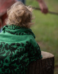 Ein Kleinkind stützt sich in der Natur auf einen Baumstumpf und hat sich eine grüne lilimoon Babydecke mit Blütenmuster wärmend um die Schultern gelegt.